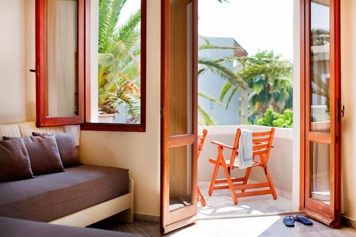 Een balkon met een houten stoel en een klein tafeltje, een bank en een raam met uitzicht op groen.