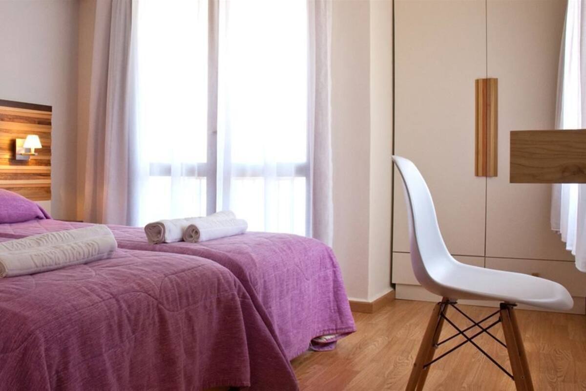 Een hotelkamer met twee bedden, een houten hoofdeinde, een witte stoel en een groot raam met gordijnen.