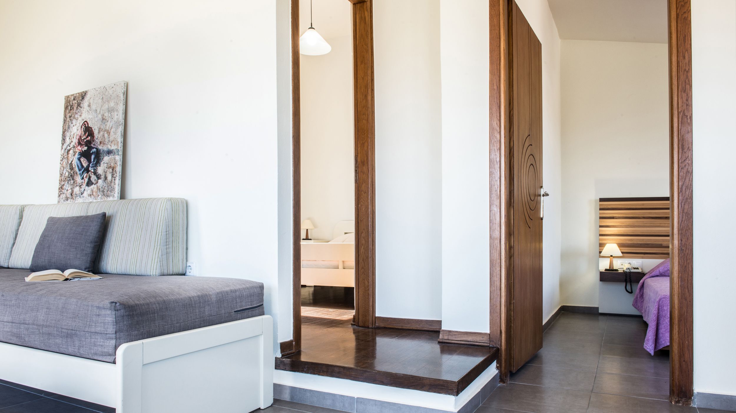 Un dormitorio moderno con una cama, una mesita de noche con lámpara y una puerta de madera que da a otra habitación.