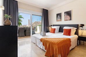 A hotel room with a large bed, a balcony with wicker furniture, and two paintings on the wall.