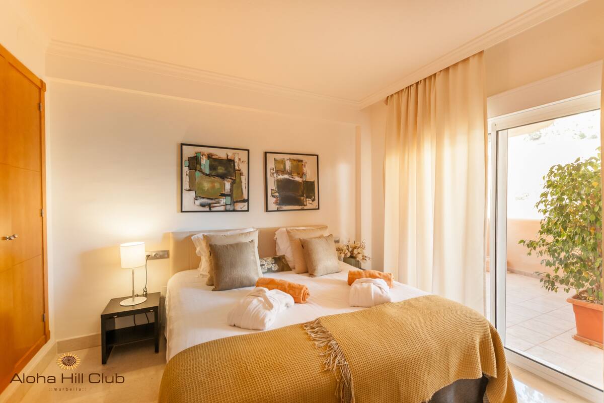 A hotel room with a bed, a nightstand, two framed artworks, a window with curtains, and a potted plant outside.