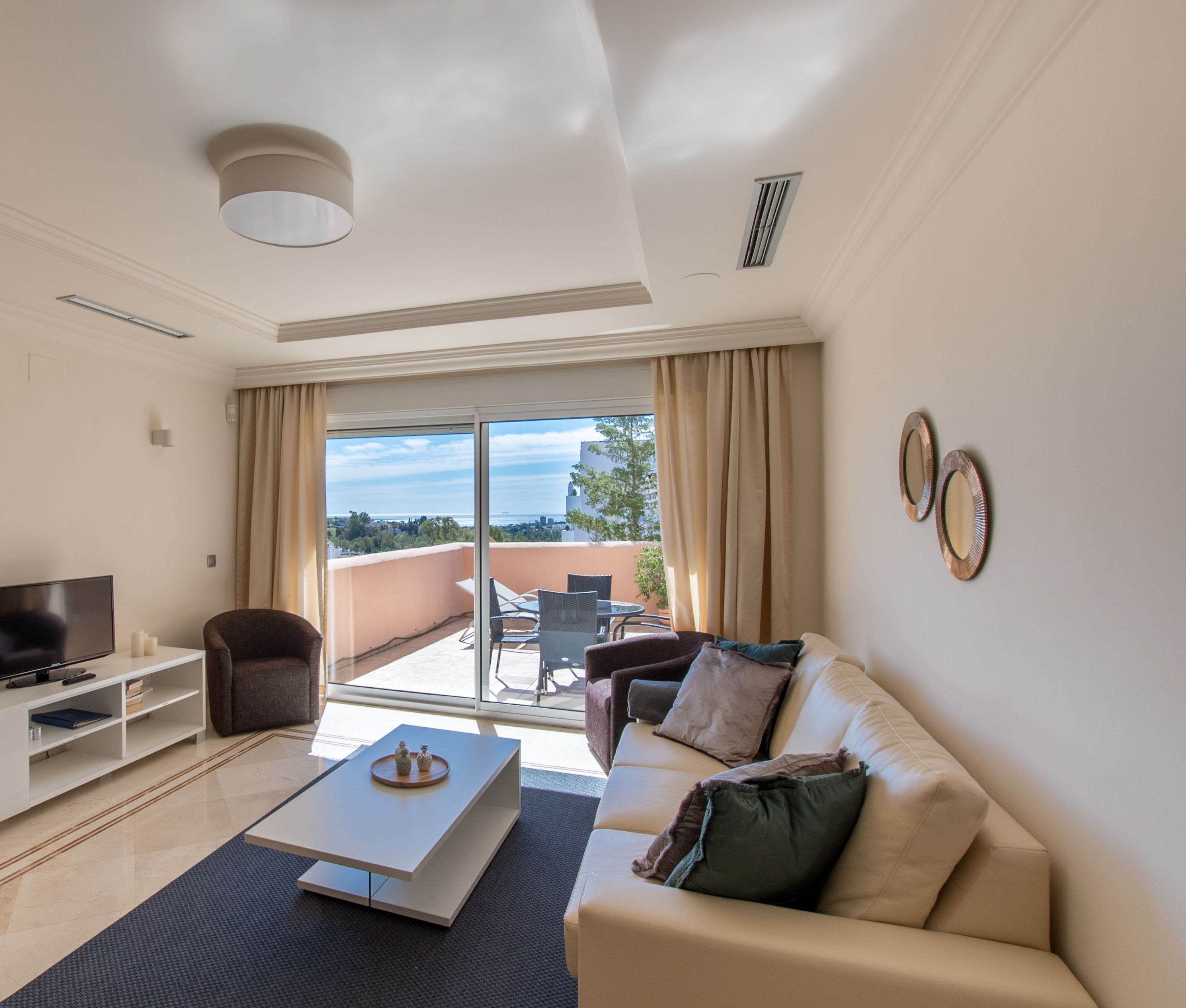 A modern living room with a sofa, coffee table, TV, and a balcony with outdoor seating.