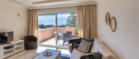 A modern living room with a sofa, coffee table, TV, and a balcony with outdoor seating.