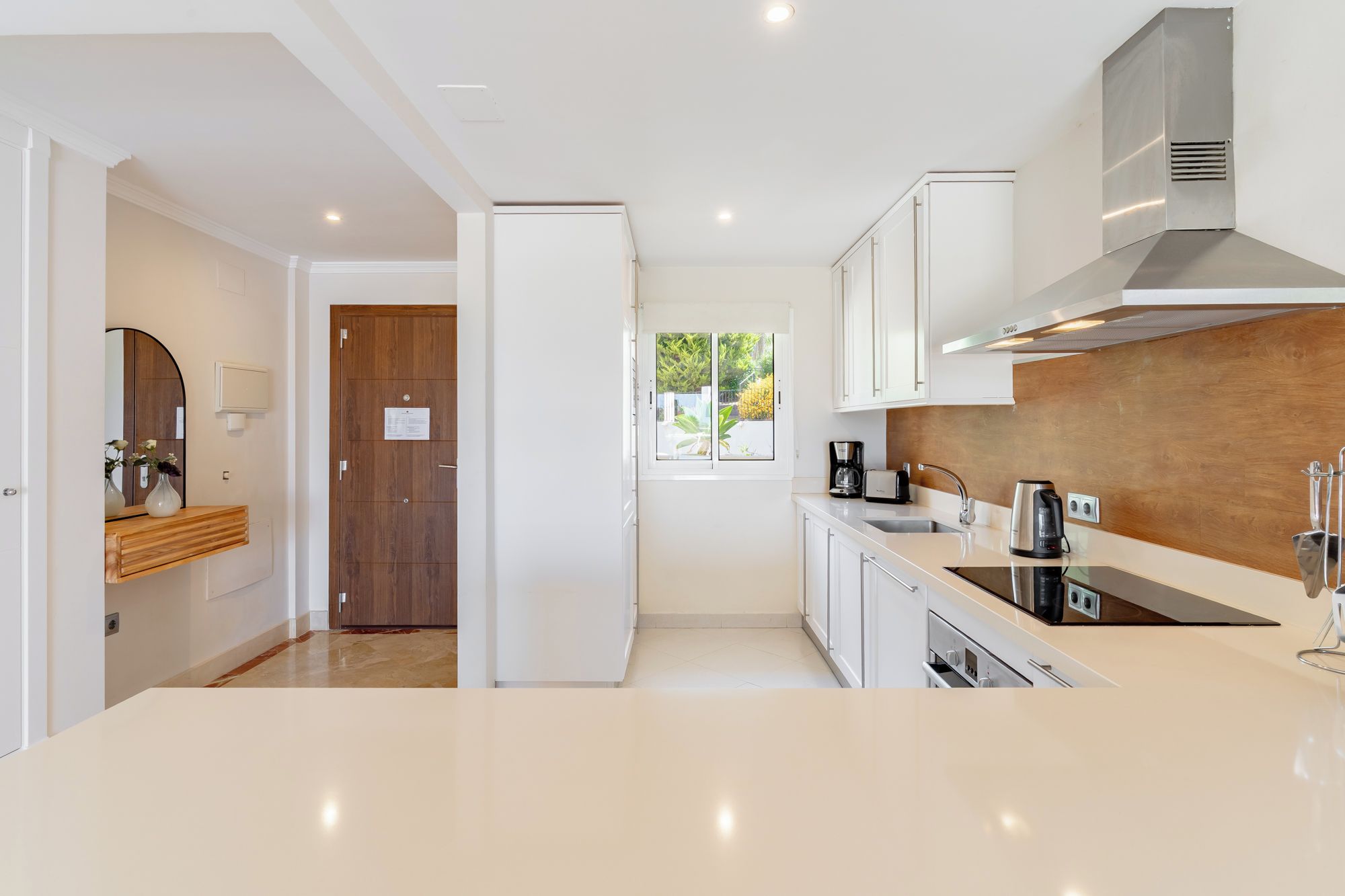 A modern kitchen with white cabinetry, a stainless steel range hood, and a window with a view of greenery.