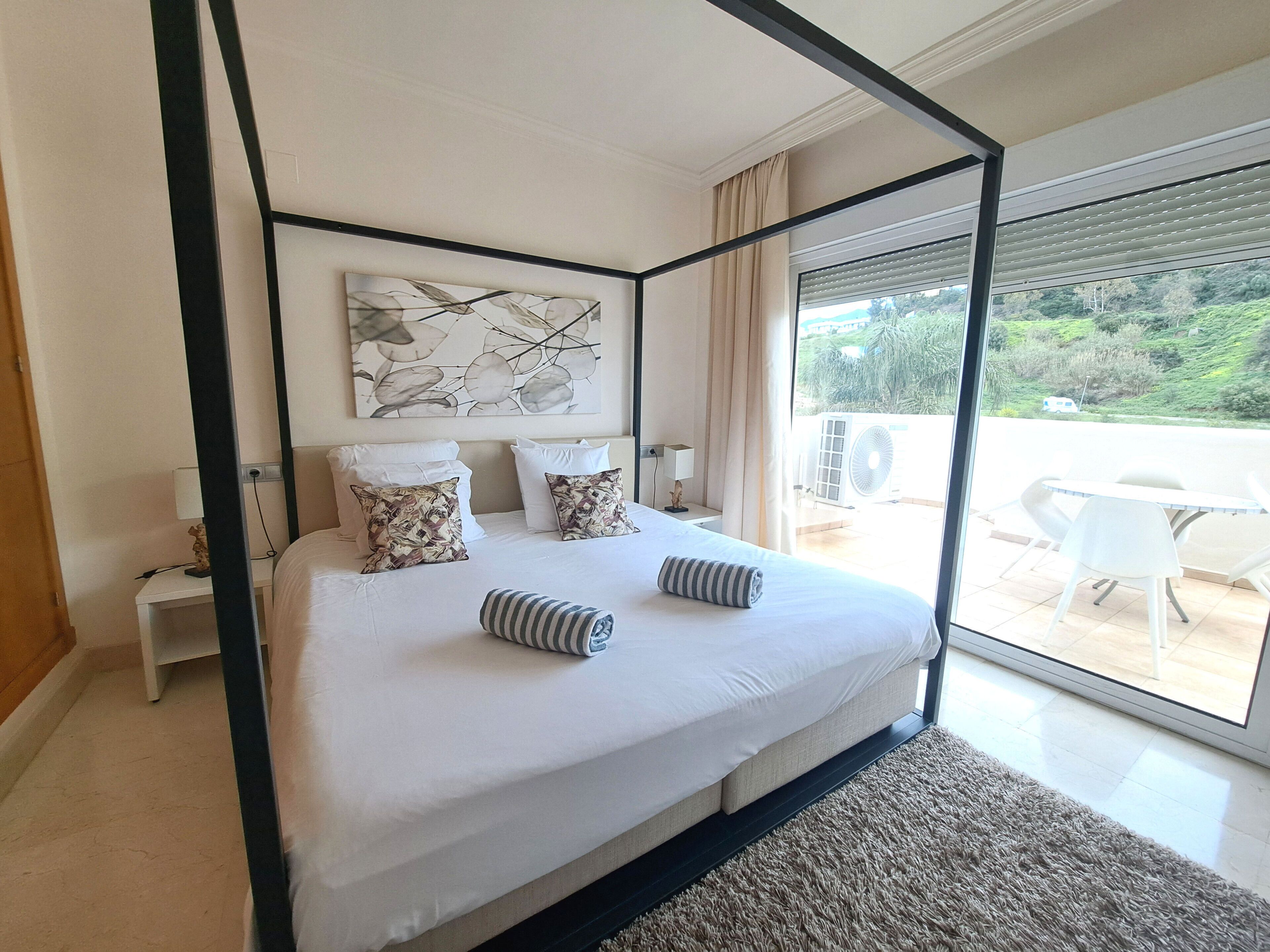A four-poster bed with white linens and patterned pillows, a large abstract painting above, and a view of greenery through a sliding glass door.
