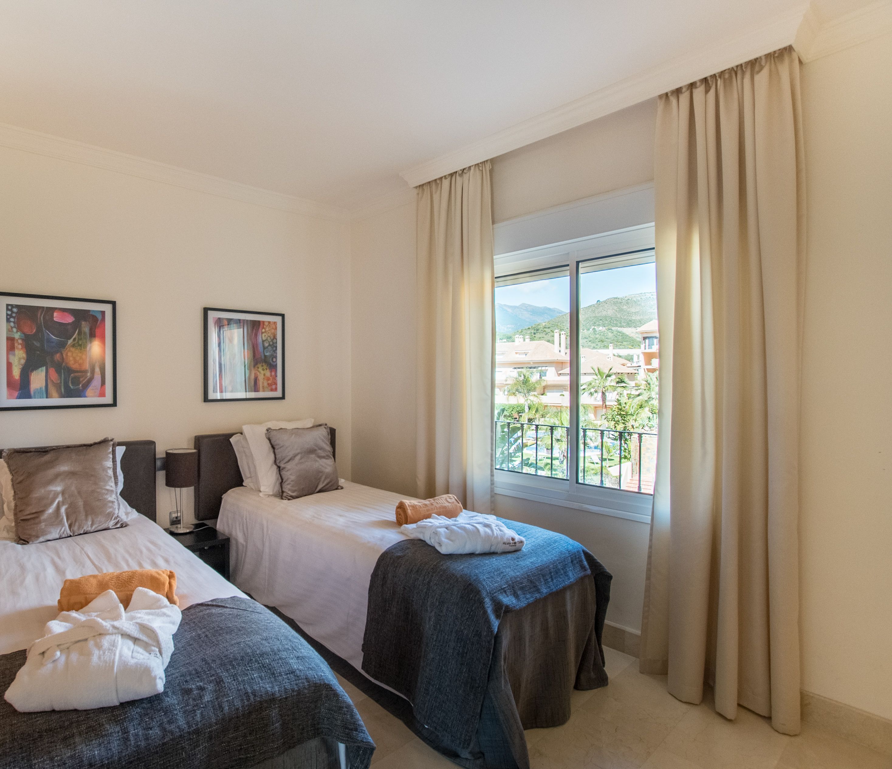 A hotel room with two beds, a large window with curtains, and artwork on the walls.