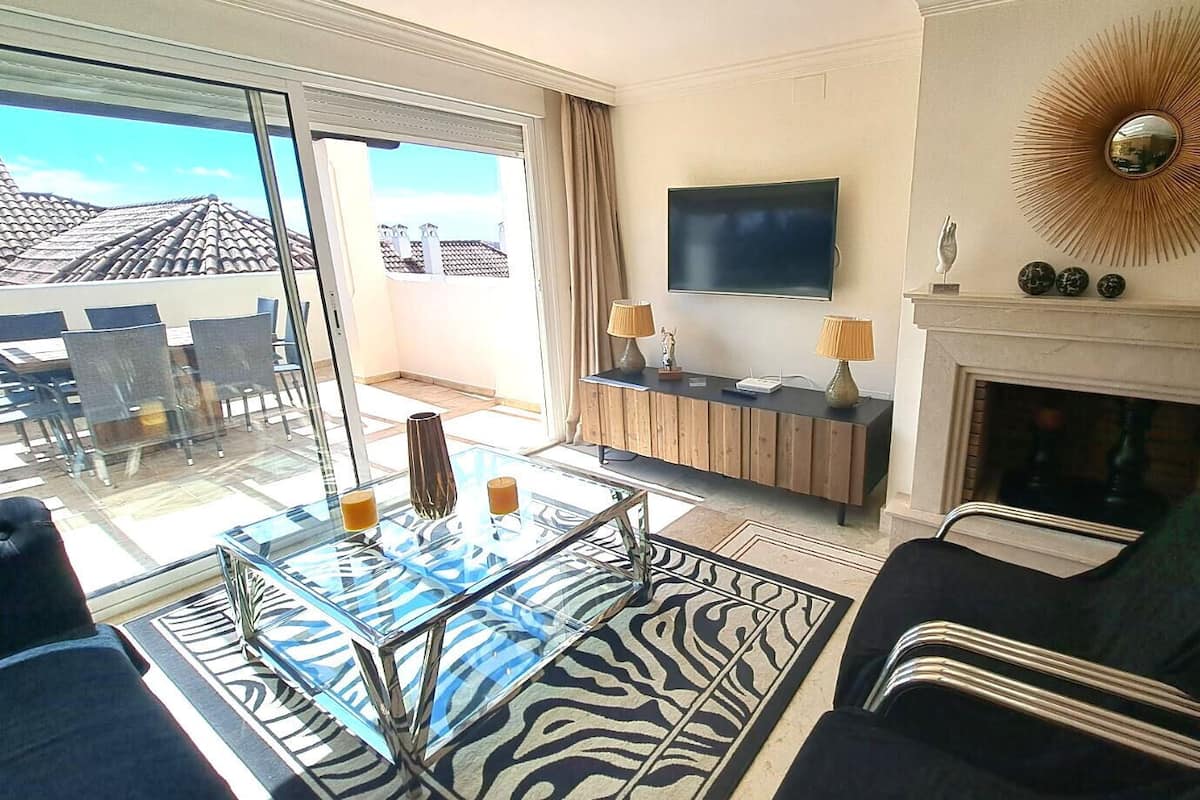 A modern living room with a glass coffee table, a fireplace, a flat-screen TV, and a view of the cityscape through large windows.