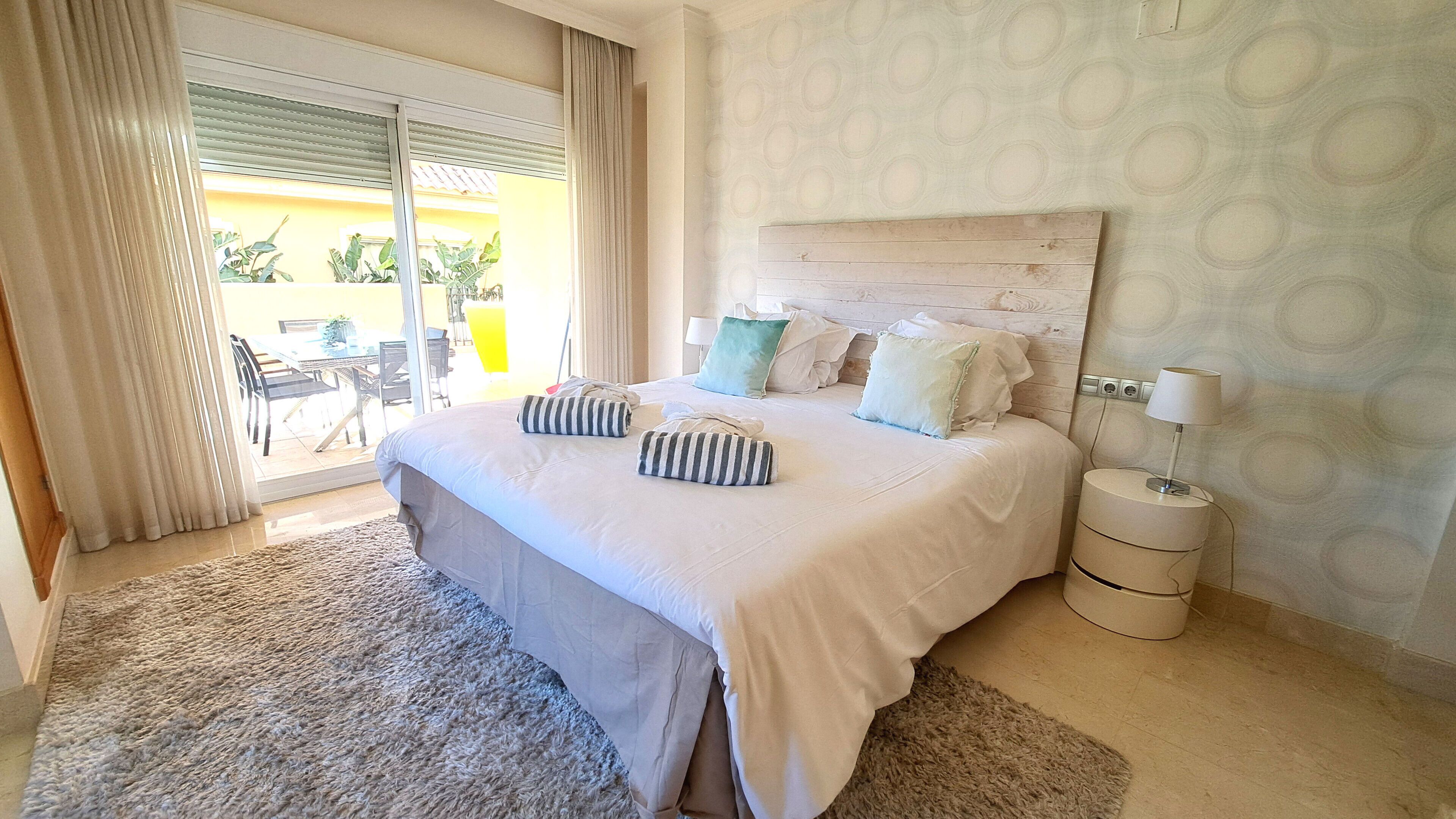 A bedroom with a large bed, a nightstand, a lamp, and a view of the outdoors through a sliding door.