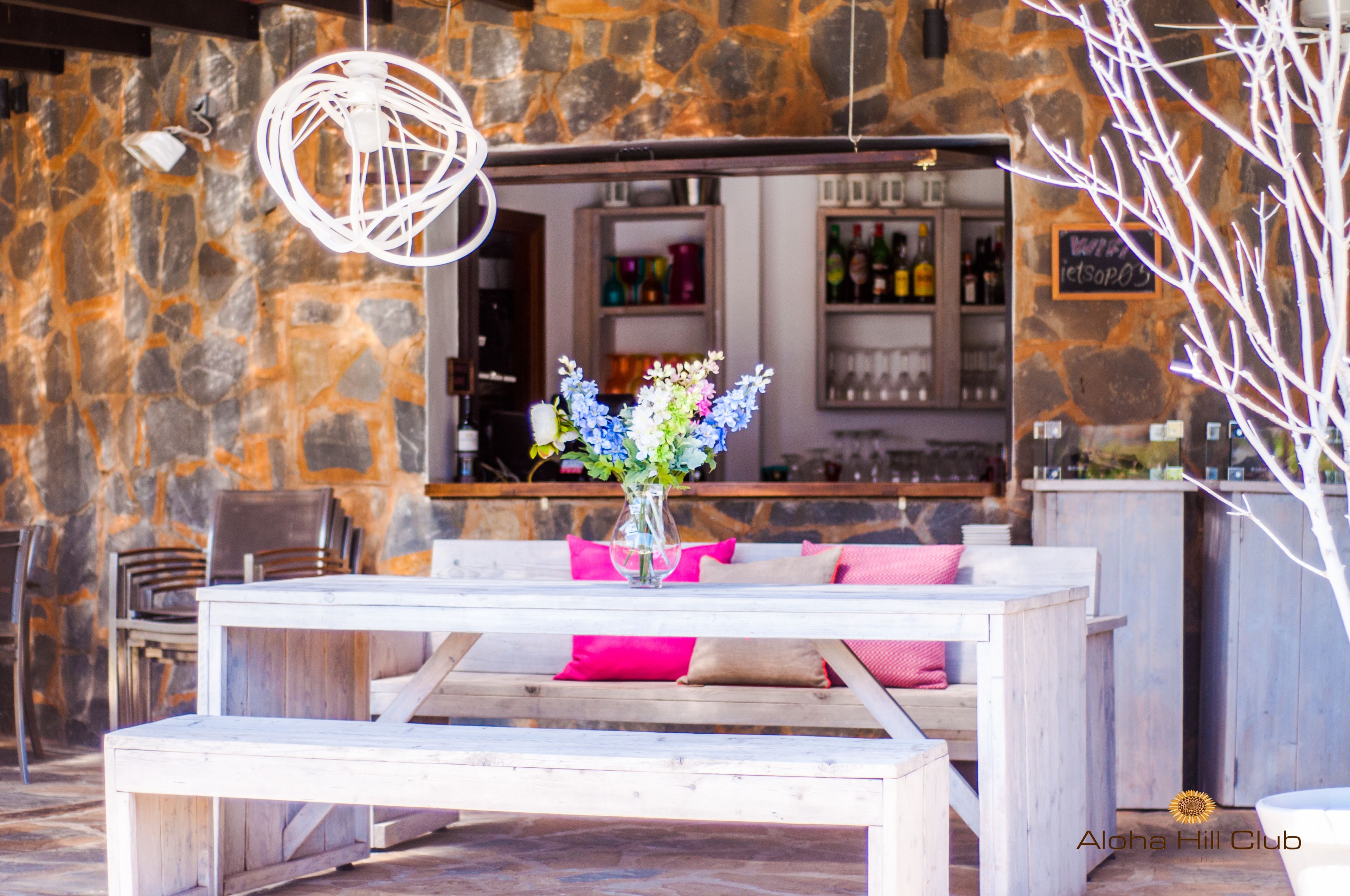 Un área de estar al aire libre con un fondo de pared de piedra, una mesa de madera con un florero y una barra en el fondo.