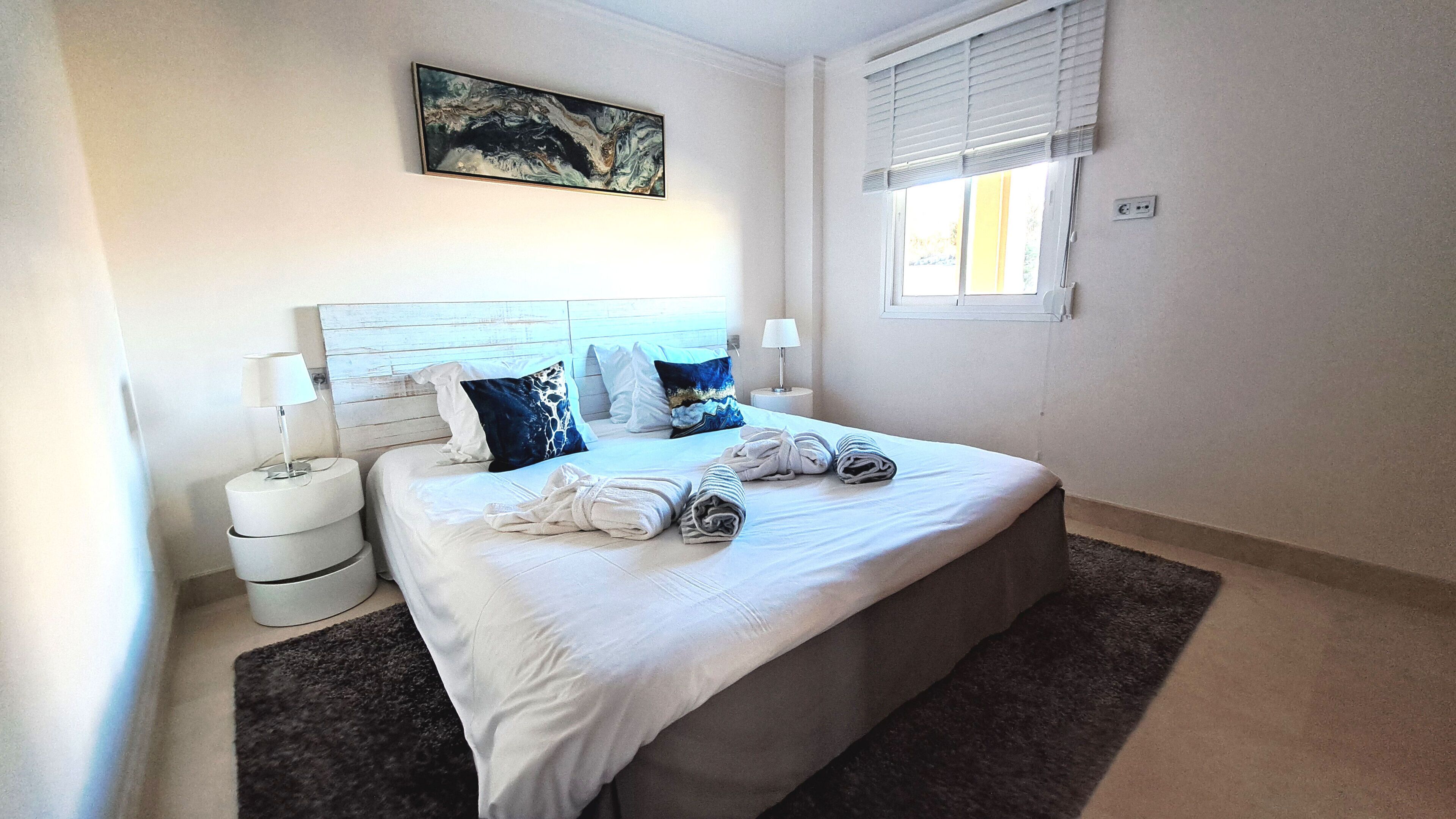 Una cama bien hecha con almohadas, una lámpara de noche y un cuadro en la pared.