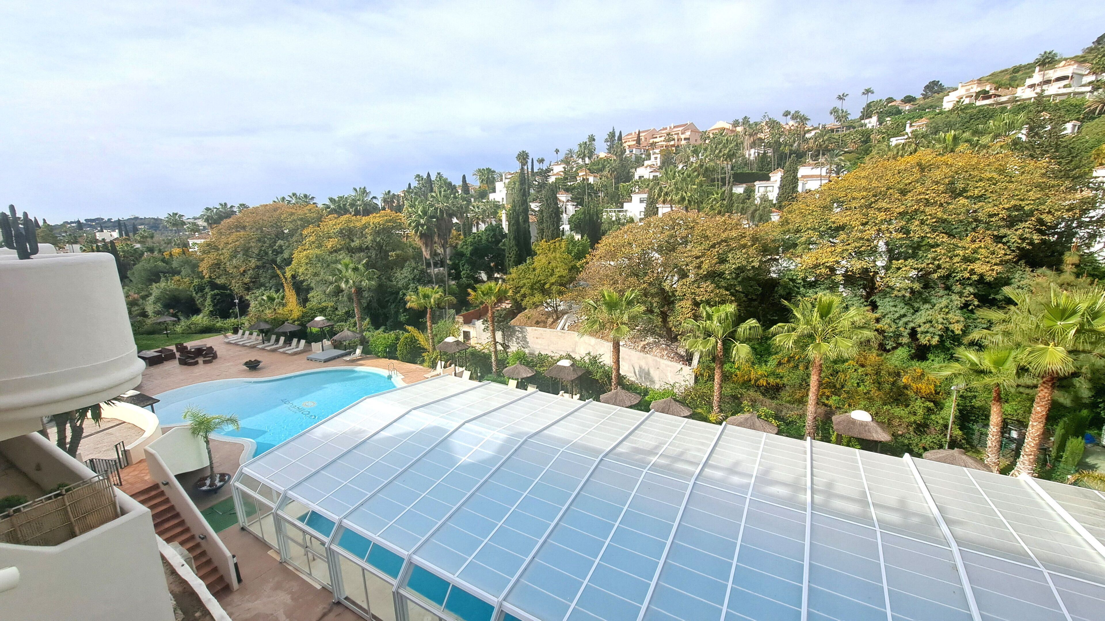 A rooftop pool with a glass enclosure, surrounded by palm trees and a view of a hillside residential area.