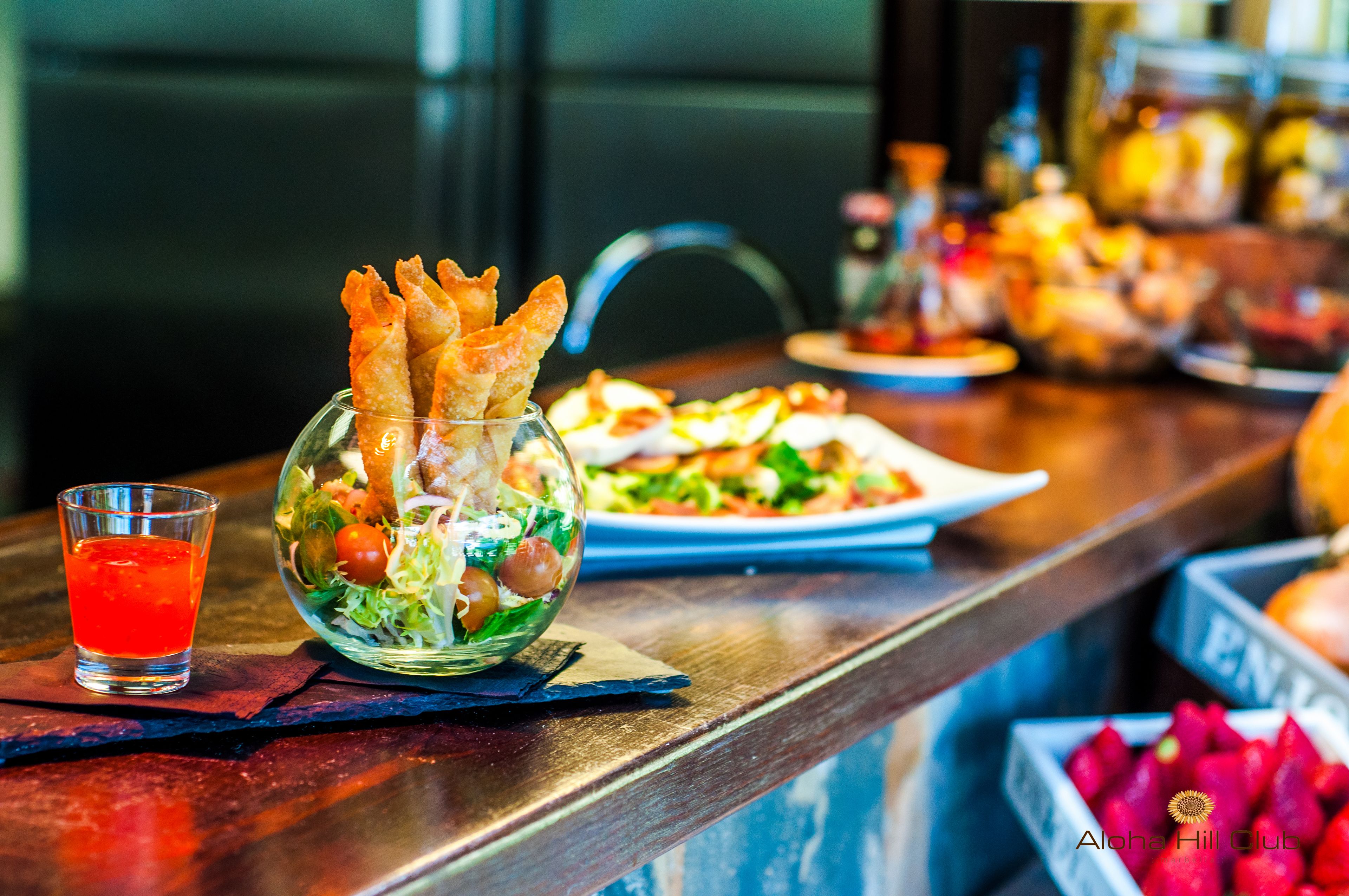 Una zona de bar con una copa de bebida roja, una ensalada en un bol de cristal y diversos platos sobre la barra.