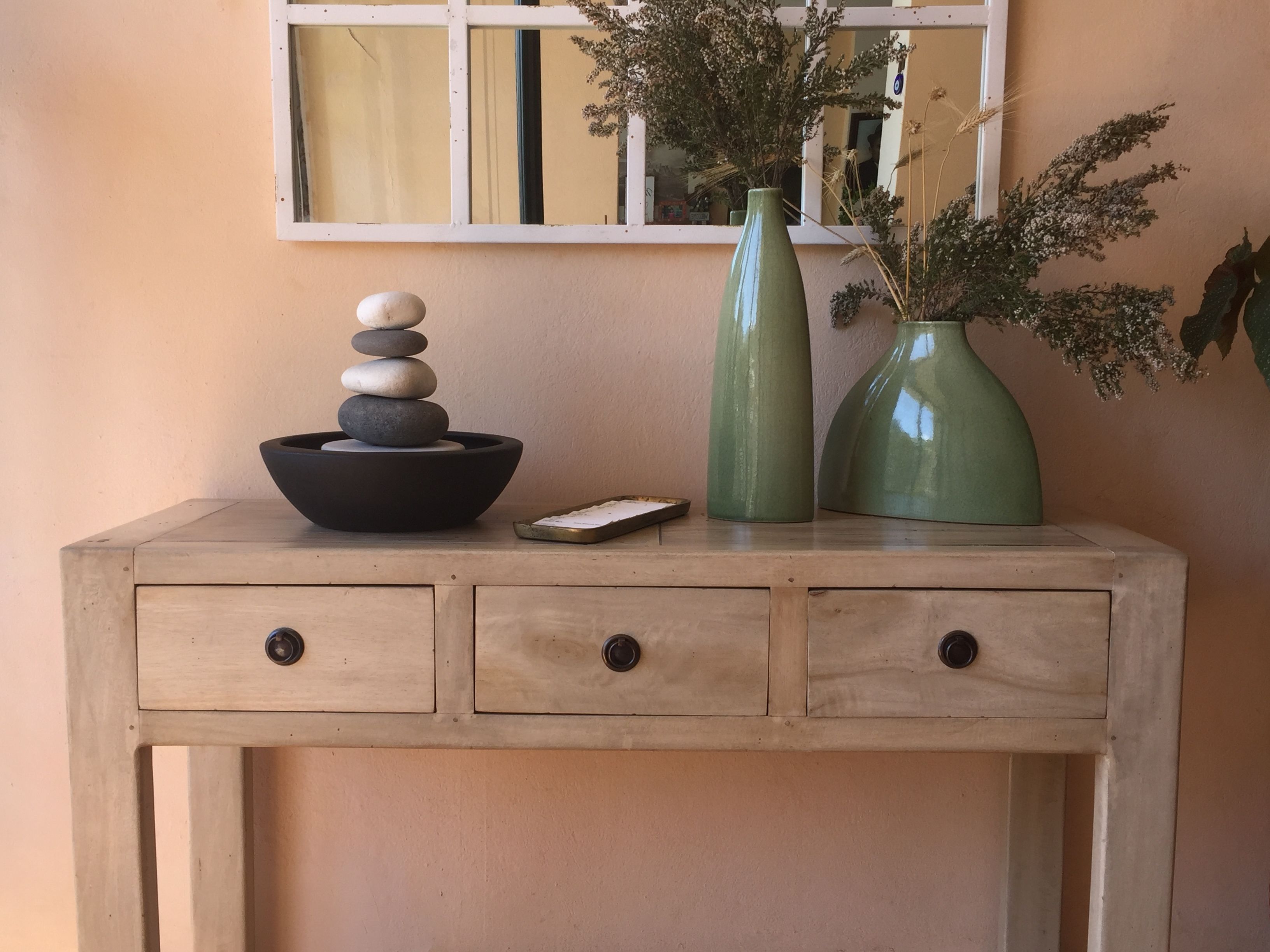 Een houten console tafel met drie lades, een schaal vol stenen, een groene vaas met gedroogde bloemen, en een klein rechthoekig dienblad.