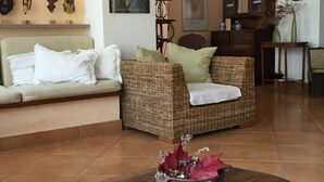 Un salon comprenant un canapé, deux fauteuils, une table basse avec des livres, et un mur orné d’un tableau encadré et d’objets décoratifs.