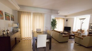 A modern living room with a dining area, beige sofas, a TV, and a large window with curtains.