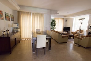 A modern living room with a dining area, beige sofas, a TV, and a large window with curtains.