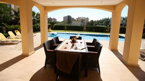 A covered outdoor dining area with a table set for two, overlooking a swimming pool and a landscaped garden.