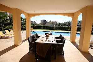 A covered outdoor dining area with a table set for two, overlooking a swimming pool and a landscaped garden.