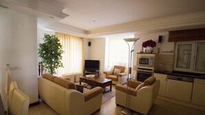 A living room with beige sofas, a coffee table, a TV, a plant, and a kitchen area with a microwave and oven.