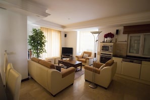 A living room with beige sofas, a coffee table, a TV, a plant, and a kitchen area with a microwave and oven.