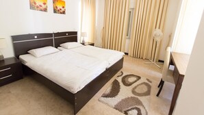 A hotel room with a double bed, bedside tables, a desk, a chair, a standing fan, and two paintings on the wall.