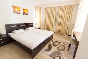 A hotel room with a double bed, bedside tables, a desk, a chair, a standing fan, and two paintings on the wall.