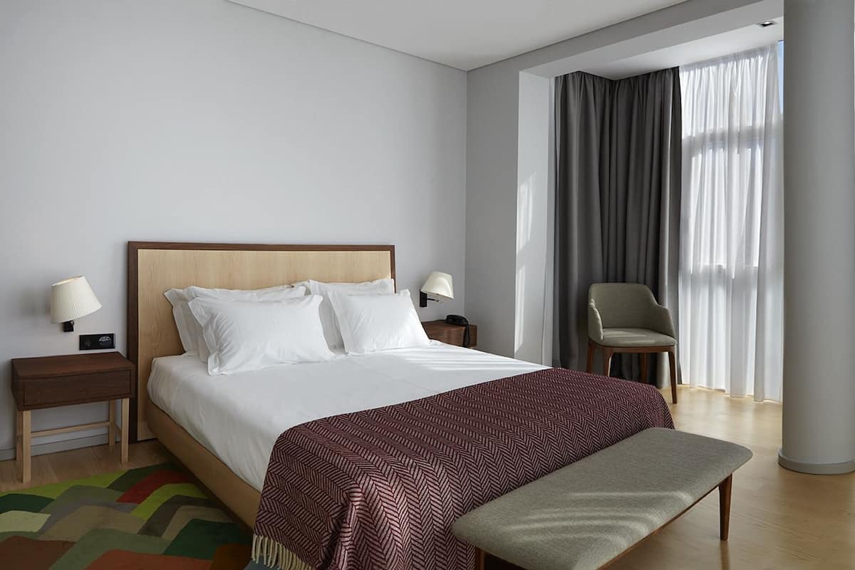 A hotel room with a large bed, two bedside lamps, a chair, and a view of the outdoors through sheer curtains.