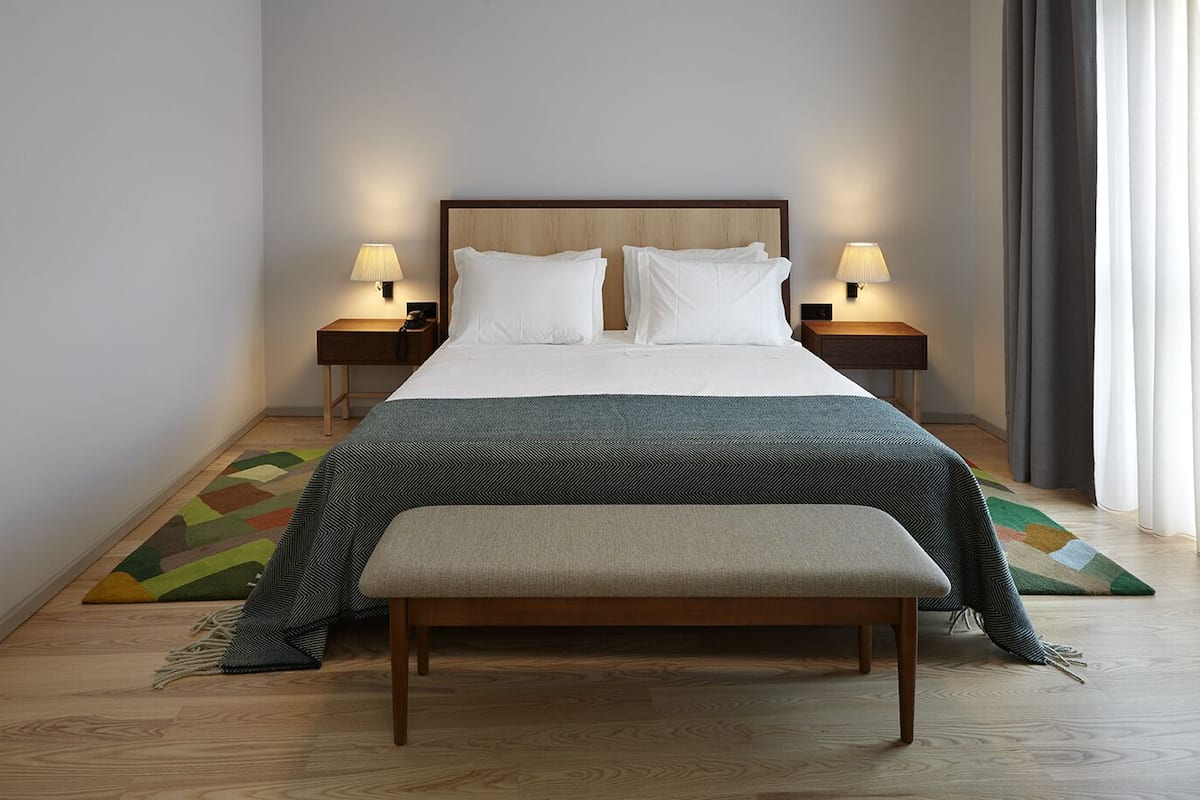 A neatly arranged bedroom with a wooden headboard, two bedside lamps, a bench, and a colorful rug.