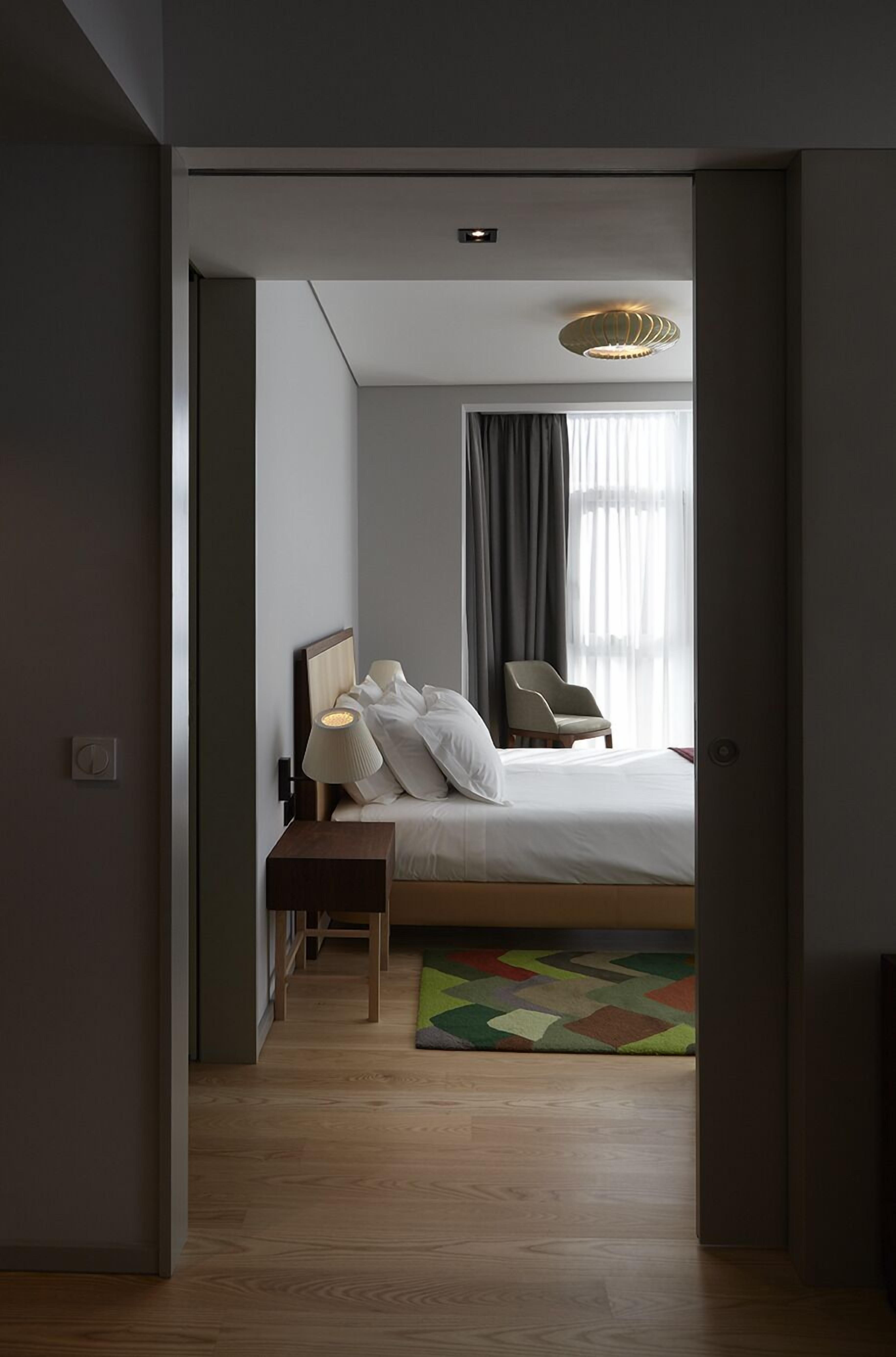 A modern hotel room with a neatly made bed, a chair, a lamp, and a colorful rug.
