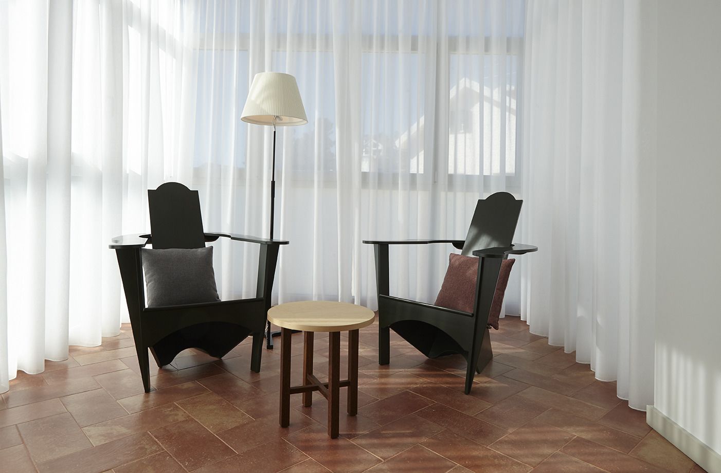 Two black chairs with brown cushions, a small round table, and a floor lamp in a room with wooden flooring and sheer white curtains.