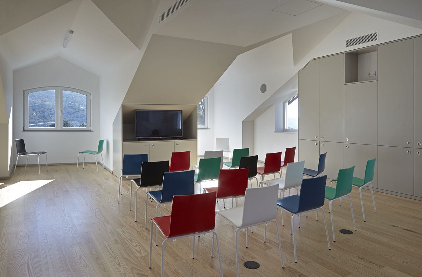A spacious room with a sloped ceiling, wooden flooring, and a large window.