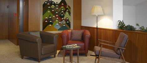 A lobby with a wooden floor, a red armchair, a brown armchair, a brown sofa, a small table with magazines, and a lamp.