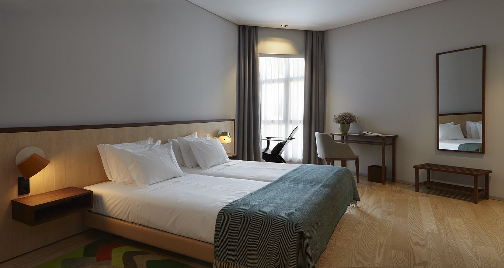Ein Hotelzimmer mit Bett, Nachttisch, Lampe, Sessel, Schreibtisch und Spiegel.