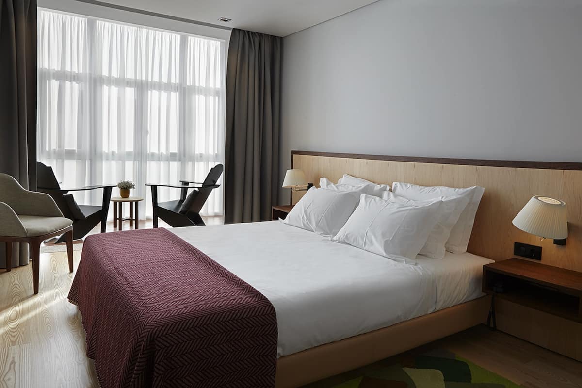 A hotel room with a large bed, a desk, two chairs, a lamp, and a window with curtains.