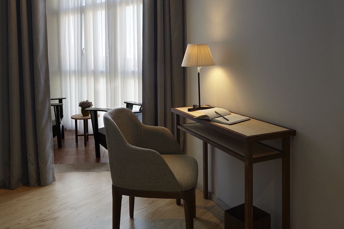 A modern room with a desk, chair, lamp, and a view through sheer curtains.