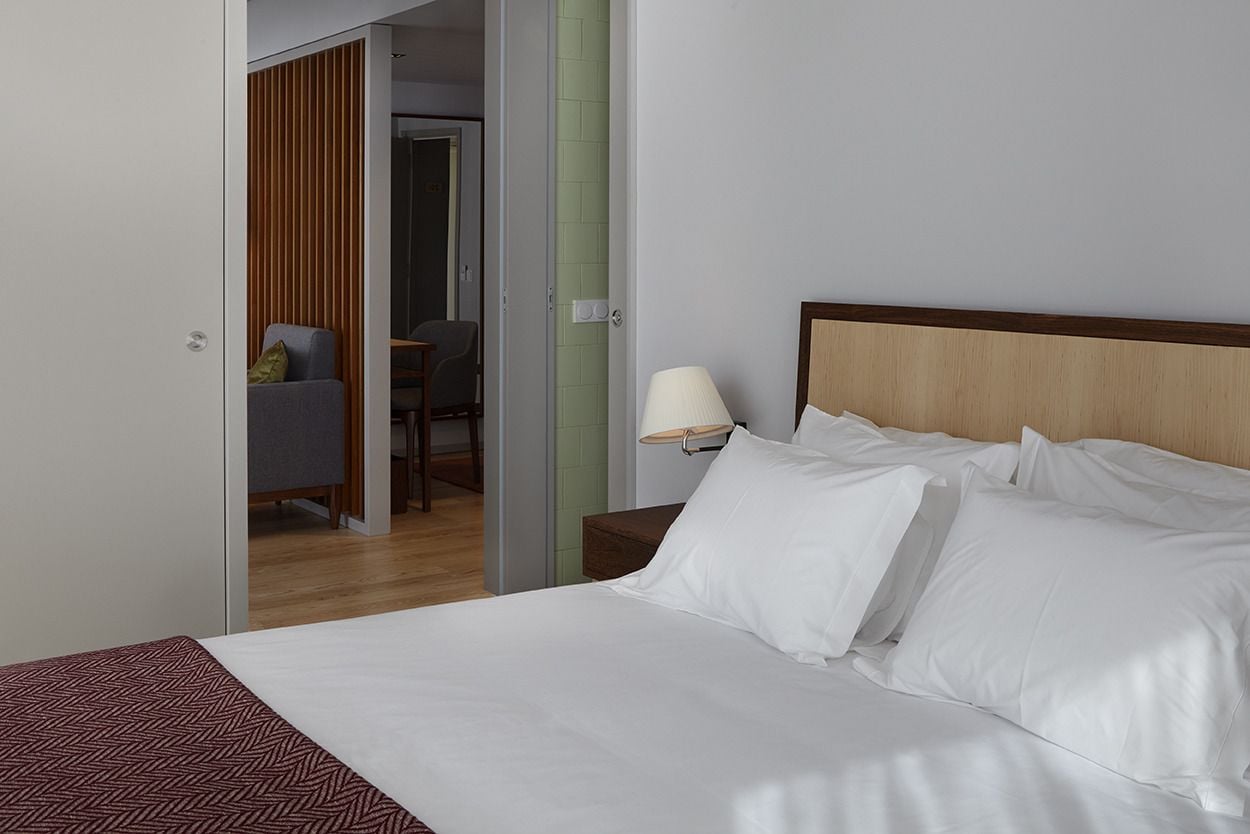 Habitación de hotel con una cama, una mesita de noche, una lámpara y vistas a un comedor a través de una puerta abierta.