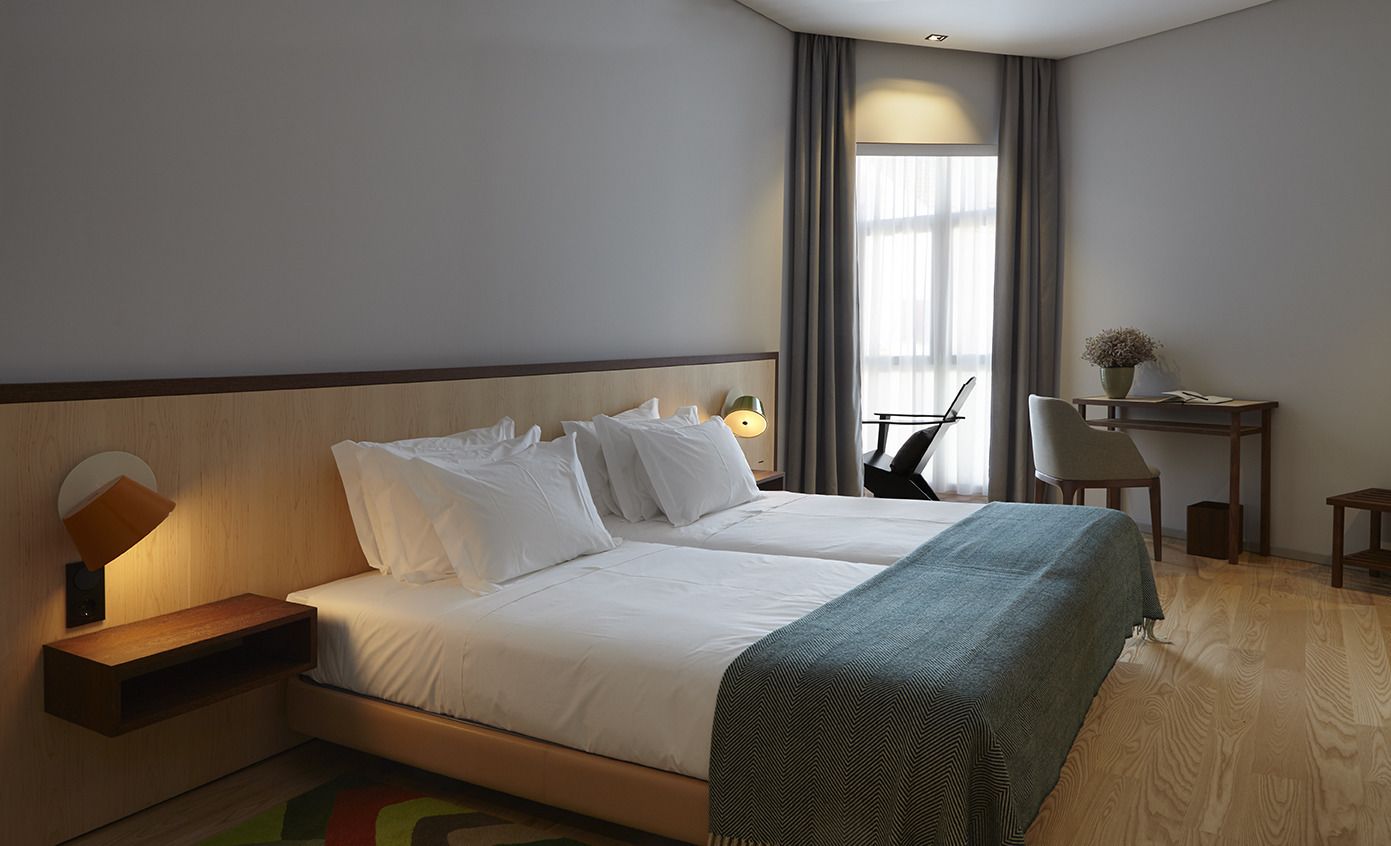 A hotel room with a double bed, a desk with a chair, a lamp, and a window with curtains.