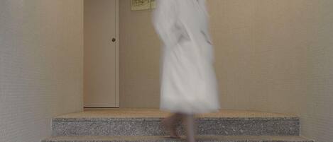 A person in a white robe descending a set of stone steps in a building with a plain wall and a door with a sign.