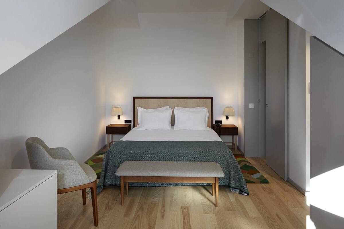 A modern hotel room with a large bed, two bedside tables with lamps, a chair, and a bench.