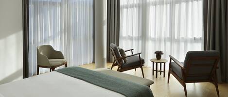 A hotel room with a large bed, two armchairs, a small table with a plant, and sheer curtains.