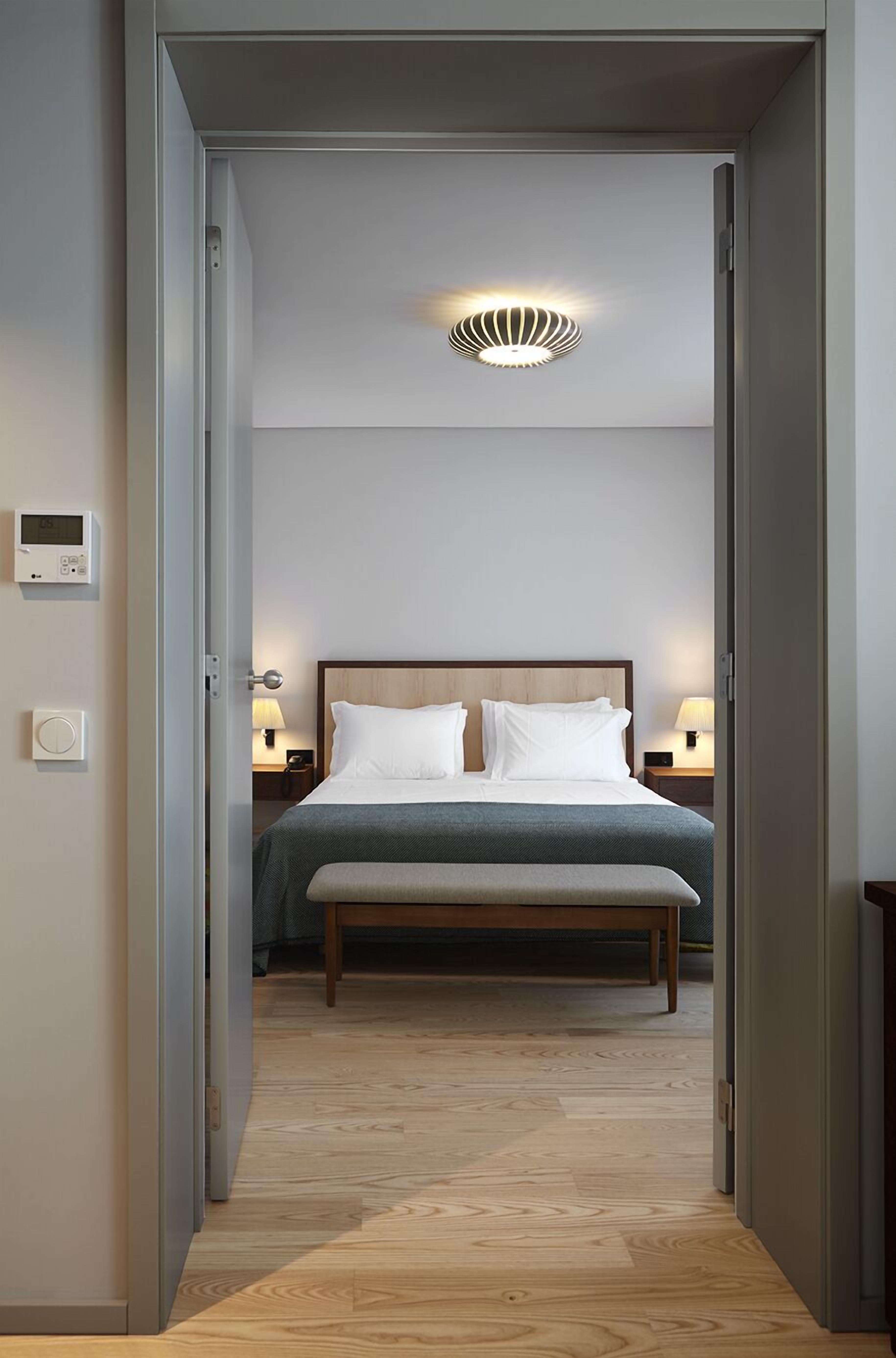 A hotel room with a bed, bedside lamps, a thermostat, and a wall-mounted light fixture.