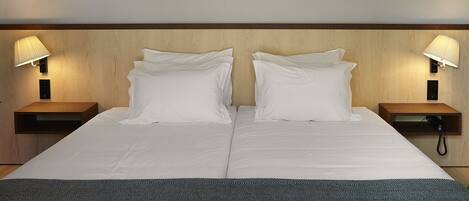 Cama doble con dos almohadas blancas y una manta gris con estampado. A ambos lados de la cama hay dos mesitas de noche con lámparas.