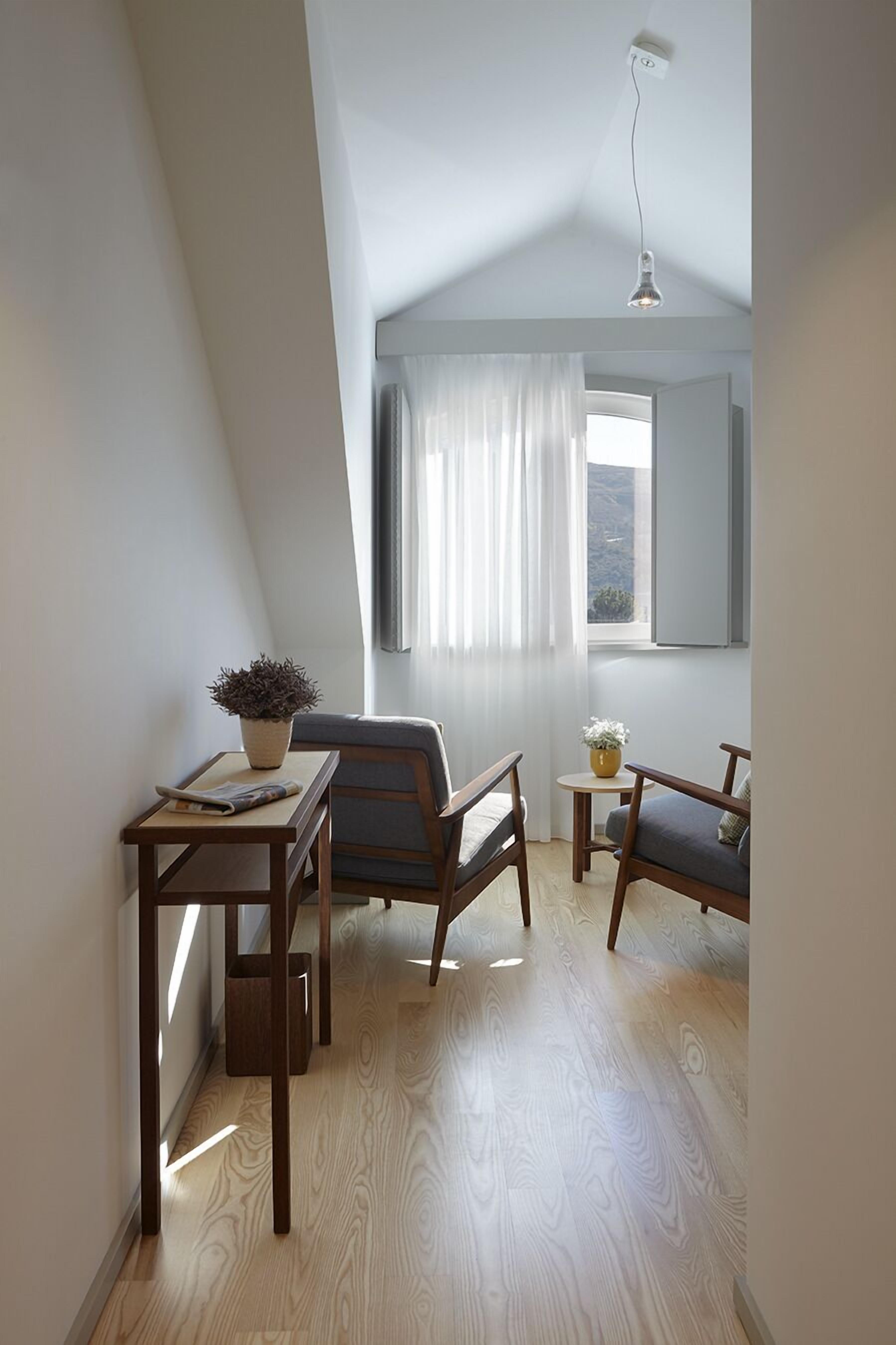 A cozy room with a sloped ceiling, a small table with a plant, two chairs, a window with curtains, and a view of the outdoors.
