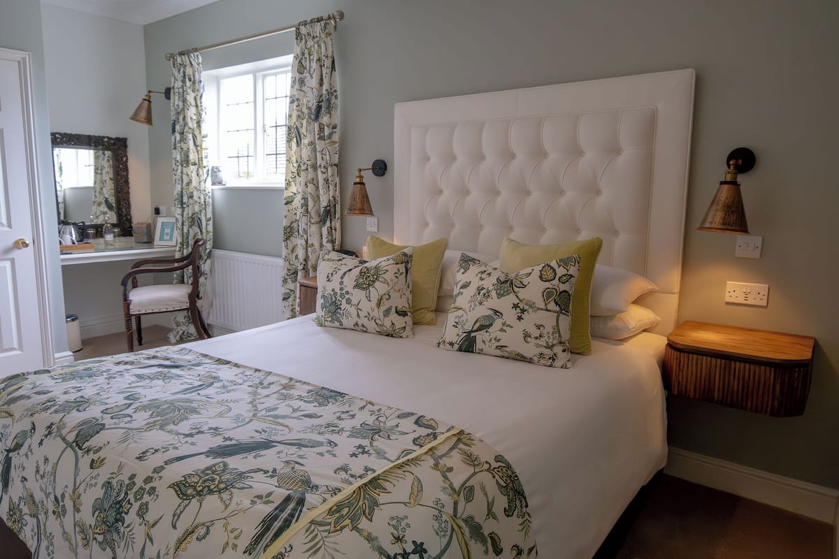 A bedroom with a large bed, floral bedding, a wooden nightstand, and a desk with a chair.