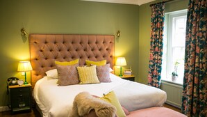 A bedroom with a tufted headboard, a large bed with multiple pillows, bedside lamps, a nightstand, and a window with floral curtains. - The Stable Courtyard Bedrooms at Leeds Castle (Maidstone)