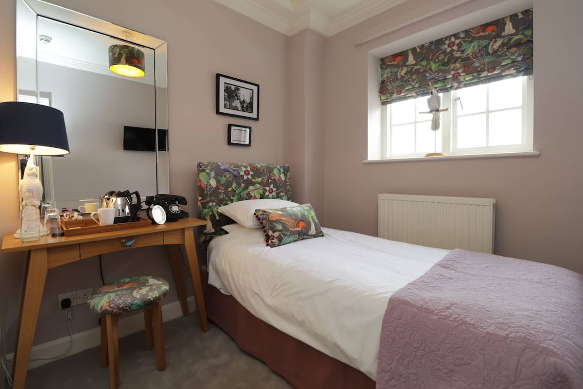 A bedroom with a bed, a desk with a lamp, a chair, a window with a floral valance, and a mirror.