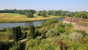 A riverside garden with a pathway, lush greenery, and a building with a brick facade. - The Stable Courtyard Bedrooms at Leeds Castle (Maidstone)