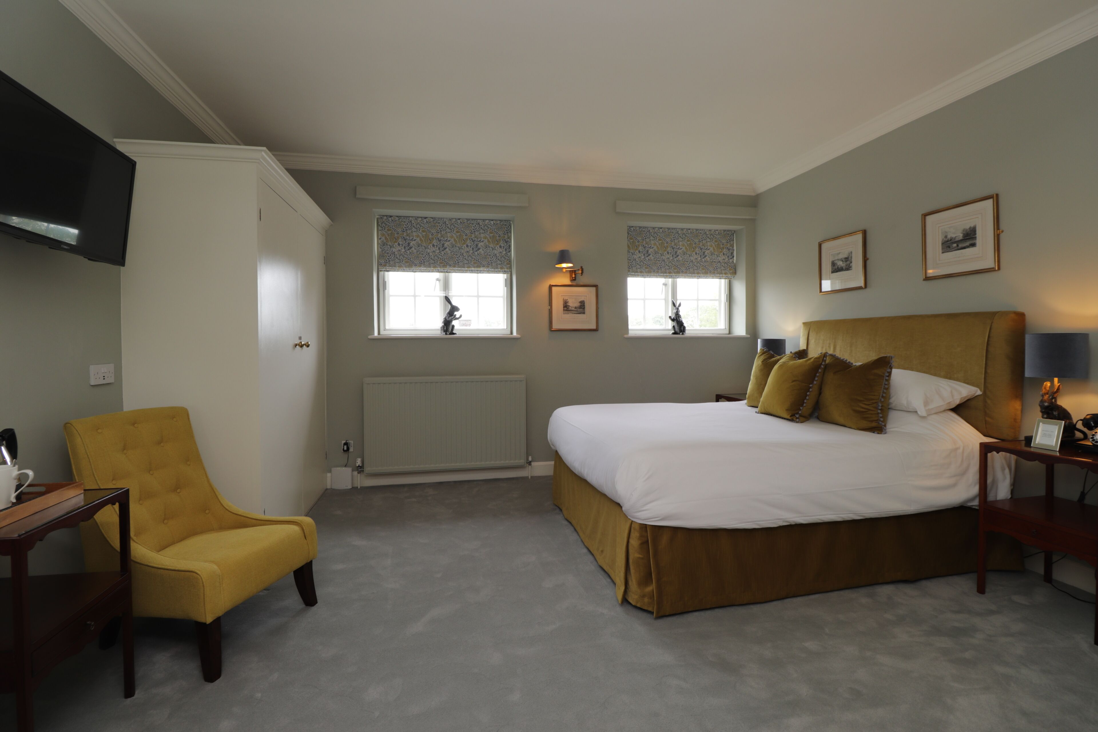 A bedroom with a bed, a yellow armchair, two windows with blinds, a flat-screen TV, and framed pictures on the wall.