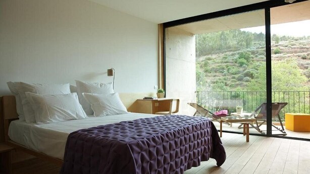 A bedroom with a large bed, a balcony with a view of the countryside, and a small table with chairs.