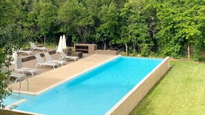 A rectangular swimming pool with a concrete edge, surrounded by a grassy area and a fence. There are lounge chairs and umbrellas near the pool.